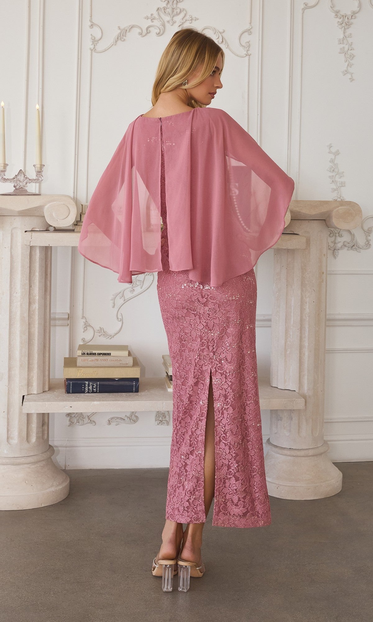 A woman in the Amoris T906 long prom dress, featuring pink sequin lace and a chiffon cape overlay, stands facing away in a room with white walls, a stone console table, candles, and stacked books.